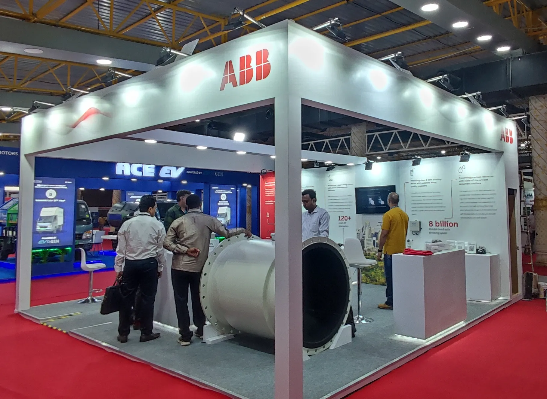 custom open island exhibition stall fabricated for ABB featuring heavy flow equipment displays, illuminated acrylic fascia, and white reception desk in Bangalore