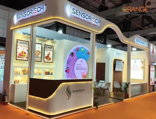 Custom Exhibition Stall Fabrication for Sensorisch at Fi India Mumbai Custom exhibition stall fabricated by Orange Stall Fabrication for Sensorisch Formulation Labs at Fi India Mumbai featuring curved backlit fascias, warm ambient LED lighting, and product discovery displays